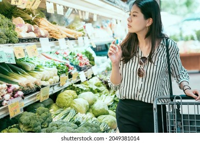 Side Portrait Of Asian Female Is Looking At The Vegetables And Thinking What To Buy While Doing Grocery Shopping At The Produce Section In A Supermarket.