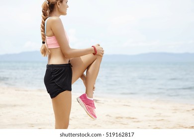 Side portrait of attractive sporty woman with long braid stretching on beach against blue sea background. Blonde female jogger in sportswear warming up her legs before morning run at the ocean