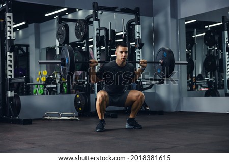 A side portrait of a bodybuilder in grey doing squats using a barbell in a gym to train his legs and back. Powerful fitness workout