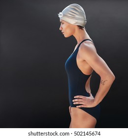 Side Portrait Of Confident Young Female Swimmer On Black Background. Young Woman In Swimsuit Standing With Her Hands On Hips.