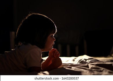 Side portrait of cute little child praying on bed in dark bedroom with backlight. Bedtime. Low key, backlit. Profile silhouette 
