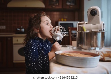 Side Portrait Cute Little Girl Making Funny Faces While Licking Whisk With Leftovers Of Whipped Cheese Cream In Steel Bowl At Home Kitchen, Standing Near A Plate With Freshly Made Tiramisu Cake