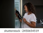 Side portrait of a dipsomaniac, alcoholic drunk young woman standing at the refrigerator in the home kitchen, choosing a bottle of wine in the morning. Alcoholism, mental health and social problems