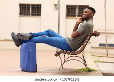 Side Portrait Of Happy African American Man Sitting Outside With Suitcase And Talking On Cell Phone