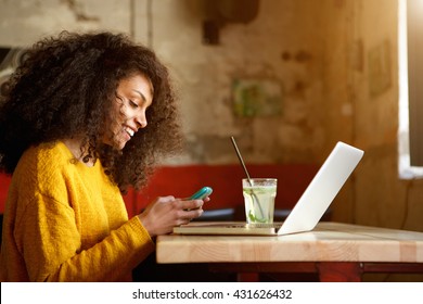 Side Portrait Of Happy Young African Woman In A Coffee Shop Using Mobile Phone