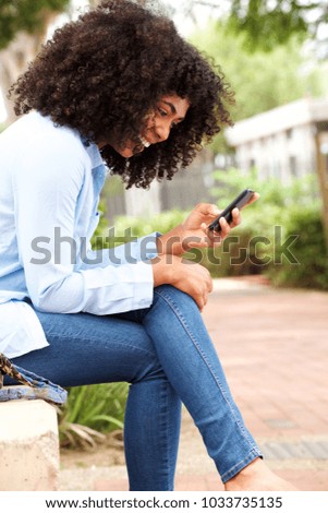 Side portrait of happy young african american woman sitting outdoors and using cell phone