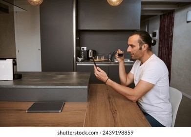 Side portrait of a relaxed young business man freelancer taking his morning coffee, reading news on his smart mobile phone, checking social media content, sitting at table in cozy home interior. - Powered by Shutterstock