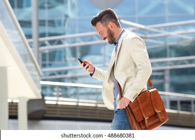 Side Portrait Of Smiling Mature Man With Bag Walking Outdoors And Reading Text Message On His Cell Phone