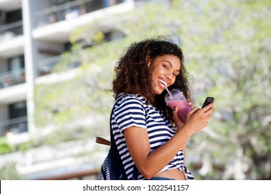 Side Portrait Of Smiling Young African American Woman Walking With Mobile Phone And Drinking Smoothie