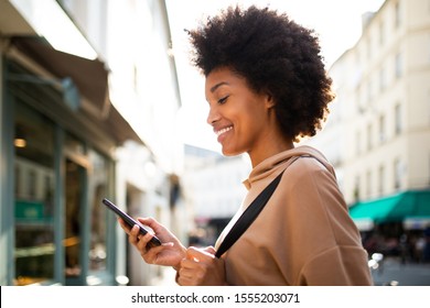 Side Portrait Of Smiling Young Black Woman Looking At Mobile Phone In City