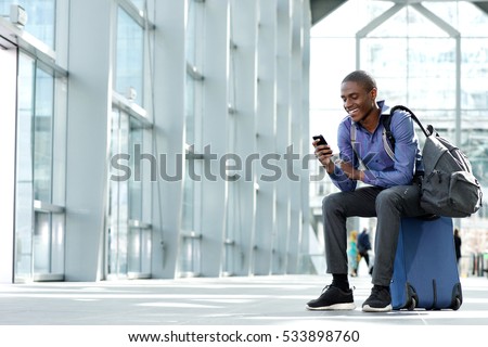 Side portrait of smiling young businessman sitting on suitcase looking at phone