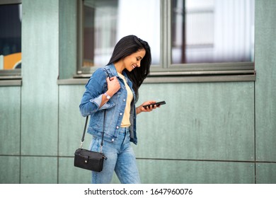 Side Portrait Of Smiling Young Hispanic Woman Walking With Mobile Phone And Purse