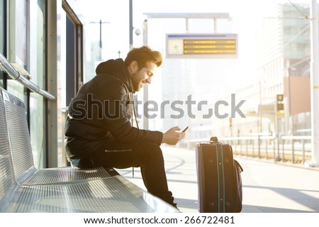 Side portrait of a smiling young man sitting with mobile phone and bag waiting for train