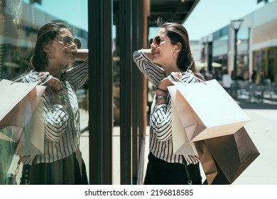 side portrait stylish asian lady holding shopping bags is flipping hair while looking at her reflection with confident smile at shop window near a mall on street - Powered by Shutterstock