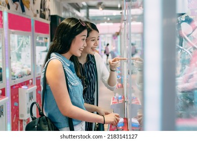side portrait two happy female friends are looking with excitement and pointing the toy in the showcase while having fun playing claw machine in a small store. - Shutterstock ID 2183141127