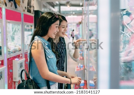 side portrait two happy female friends are looking with excitement and pointing the toy in the showcase while having fun playing claw machine in a small store.