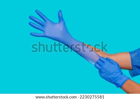 Side portrait of unrecognizable female nurse hands putting on blue medical protective sterile gloves isolated over blue background. Health care, medicine, protection, hospital concept.
