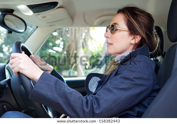 Side portrait view of a mature\
middle aged smart woman sitting on the drivers seat of a new car,\
driving through a countryside road during a sunny winter\
day.
