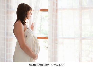 Side Portrait View Of A Young Pregnant Woman Standing By Large French Glass Doors At Home, Holding The Curtain Open And Looking At The Window Being Thoughtful And Holding Her Belly.