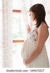 Side Portrait View Of A Young Pregnant Woman Standing By Large French Glass Doors At Home, Holding The Curtain Open And Looking At The Window Being Thoughtful And Holding Her Belly.