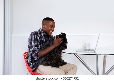 Side Portrait Of Young African Guy Sitting At A Table Playing With His Pet Dog At Home While Working On Laptop.