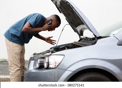 Side Portrait Of A Young African Man Having Trouble With His Broken Car Calling For Help On Cell Phone.