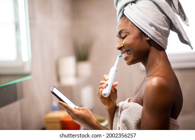 Side portrait young black woman brushing teeth and looking at mobile phone - Powered by Shutterstock