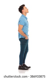 side portrait of young man in blue shirt looking up with hands in pockets while posing in isolated studio background