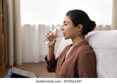 Side profile attractive young Indian woman put digital tablet on laps distracted from modern tech usage, hold glass drink still water sit on floor leaned on bed. Healthy life habit, lifestyle concept
