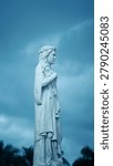 Side profile of the elegant white marble statue of Saint Philomena holding lilies, set against a dramatic, moody blue sky at the cathedral grounds in Mysore.