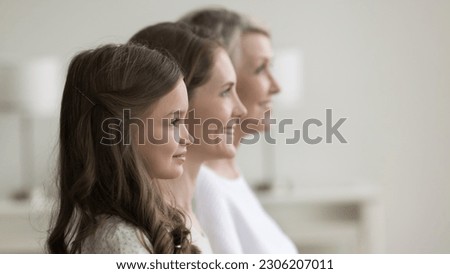 Side profile faces view of happy three generation of relatives women. Beautiful preschooler girl looking forward standing posing indoors in row with young pretty mother and senior cheerful grandmother