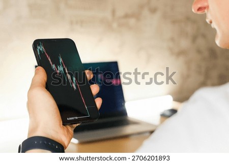 Side profile of individual checking falling financial data on smartphone beside open computer on desk