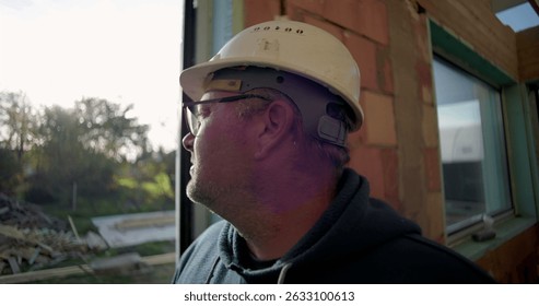 Side profile of mature construction worker in hard hat looking outdoors, standing near doorway, natural surroundings and debris in background