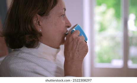 Side profile of an older woman drinking from a blue mug, savoring a hot beverage, symbolizing relaxation, comfort, and enjoying a peaceful moment in a cozy home setting - Powered by Shutterstock