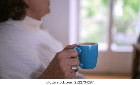 Side profile of an older woman drinking from a blue mug, savoring a hot beverage, symbolizing relaxation, comfort, and enjoying a peaceful moment in a cozy home setting - Powered by Shutterstock