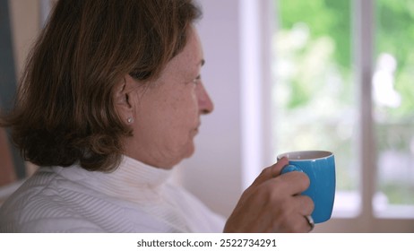 Side profile of an older woman drinking from a blue mug, savoring a hot beverage, symbolizing relaxation, comfort, and enjoying a peaceful moment in a cozy home setting - Powered by Shutterstock