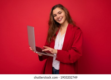 Side Profile Photo Of Charming Smiling Confident Pretty Young Lady Holding Laptop Wearing Red Cardigan And White T-shirt Isolated On Red Wall Background.