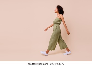 Side profile photo of cheerful nice woman with wavy hairdo dressed khaki overall white sneakers walking isolated on beige color background