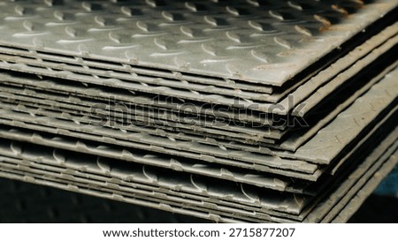 A side profile photograph of a stack of checker plates, focusing on the edges. The image shows the varying thicknesses and the rough cut of the steel sheets. The raised diamond pattern is visible on the top surfaces of the plates. This composition conveys a sense of heavy industrial activity and raw material storage.