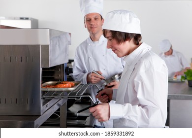 A side profile shot of a chef watching a young trainee grilling food in a kitchen.