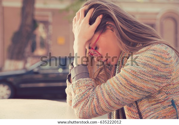 Side\
profile stressed sad young woman sitting outdoors with broken down\
car on background. City urban life style stress\
