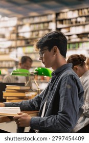 Side profile of student reading open book at quiet library table with classmates behind, focused learning, coursework and academic research for higher education, Study session.