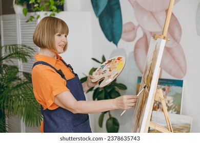 Side profile view elderly fun confident artist woman 50 years old wearing casual clothes stand near easel with painting artwork paint spend free spare time in living room indoor. Leisure hobby concept
