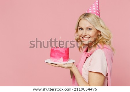 Side profile view elderly smiling cool happy woman 50s wear t-shirt birthday hat hold cake with candle look camera isolated on plain pastel pink background studio Celebration party holiday concept.