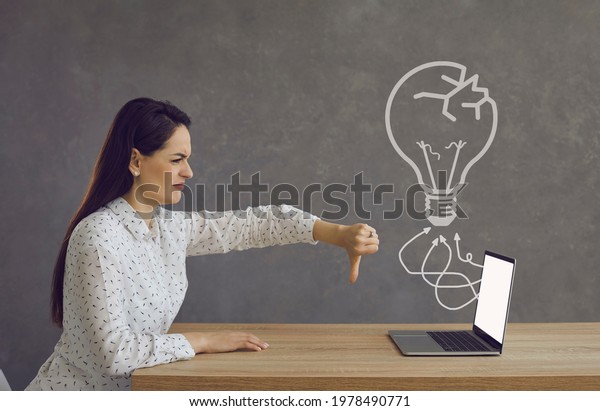 Side profile view female user at office desk\
giving thumbs down at laptop computer screen with cracked light\
bulb doodle showing employee disagreement with ineffective idea or\
bad attitude to failure