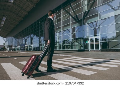 Side Profile View Full Body Young Traveler Businessman Man In Black Suit Facial Mask Walk Outside At International Airport Terminal With Suitcase Valise Crossing Road Air Flight Business Trip Concept