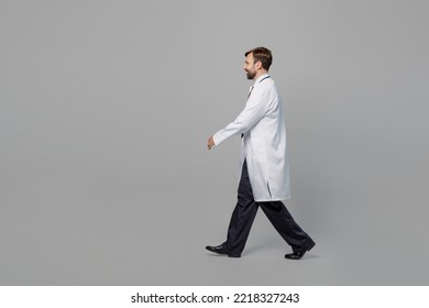 Side Profile View Full Body Male Confident Doctor Man Wears White Medical Gown Suit Work In Hospital Walking Going Isolated On Plain Grey Color Background Studio Portrait. Healthcare Medicine Concept