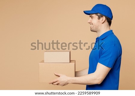 Side profile view fun delivery guy employee man wear blue cap t-shirt uniform workwear work as dealer courier hold stack cardboard blank boxes isolated on plain light beige background. Service concept