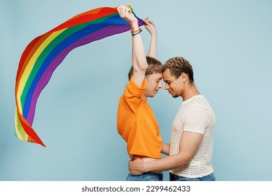 Side profile view fun young couple two gay men wearing casual clothes hold flag together isolated on pastel plain light blue color background studio portrait. Pride day june month love LGBTQ concept
