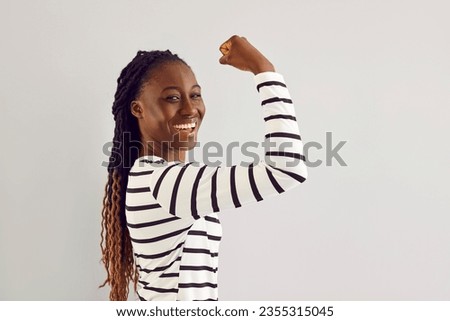 Side profile view happy joyful confident beautiful young African American woman standing on white studio background, showing her strong arm, looking at camera and smiling. Feminism, girl power concept